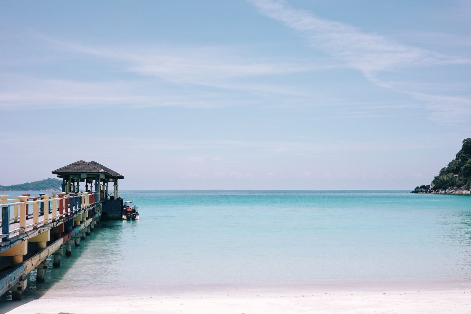 Wooden jetty reaching into calm turquoise water from a white-sand beach in the Perhentian Islands.